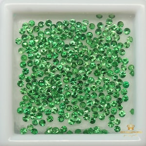 May include: A close-up view of a collection of small, round, green gemstones in a white square container. The gemstones are arranged in a random pattern and appear to be loose.