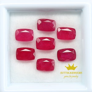 May include: Seven red ruby gemstones in a rectangular shape are displayed in a white box. The gemstones are arranged in two rows with three in the top row and four in the bottom row. The box has a white background and a white border. The text "SUTTIKARN GEMS gems for jewelry" is printed in black at the bottom of the box.