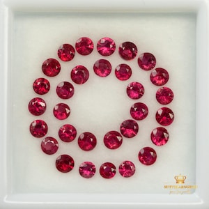 Peut inclure: Une collection de vingt-quatre rubis rouges vifs, ronds et facettés, disposés en spirale dans un présentoir carré blanc. Chaque pierre précieuse présente une taille brillant avec de multiples facettes qui reflètent la lumière, créant une teinte cramoisie.