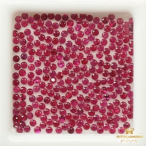 May include: A collection of small, round, faceted rubies in varying shades of red and pink, filling a white square container. The gemstones are densely packed, creating a sparkling display. The image is well-lit, highlighting the gems' brilliance.