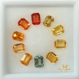 May include: A collection of emerald-cut gemstones in various colors, including orange, yellow, and a single teal stone. The gems are arranged in a circular pattern within a white square container, showcasing their facets and brilliance.