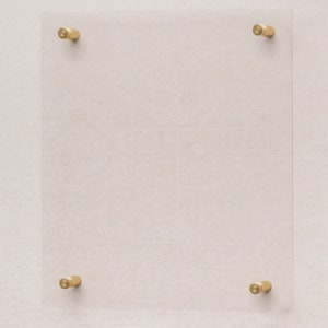 Puede incluir: Un marco de vidrio transparente con cuatro soportes de montaje de metal dorado. El marco está montado en una pared de color beige claro.