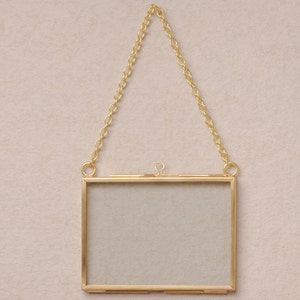 May include: A gold-toned metal picture frame with a hanging chain. The frame is rectangular and has a clear glass front.