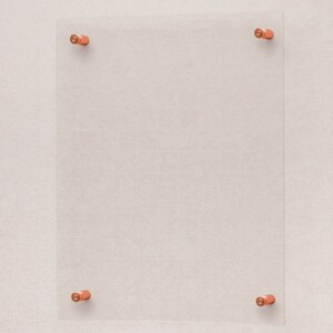 May include: A clear glass frame with four copper-colored mounting brackets. The frame is mounted on a light beige wall.