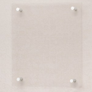 Op de afbeelding: Een transparant glazen paneel met vier zilveren metalen bevestigingsbeugels. Het paneel is gemonteerd op een lichtbeige muur.