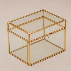 May include: A rectangular glass box with a gold-colored metal frame and a hinged lid. The box has a simple, elegant design, suitable for storing jewelry or small keepsakes. The clear glass allows visibility of the contents.