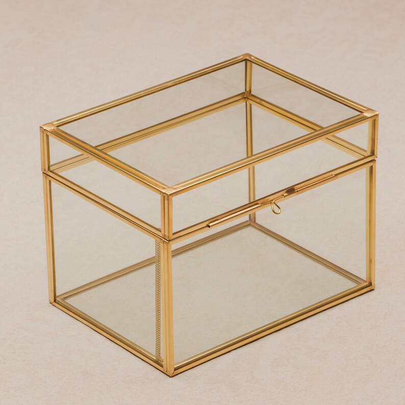 May include: A rectangular glass box with a gold-colored metal frame and a hinged lid. The box has a simple, elegant design, suitable for storing jewelry or small keepsakes. The clear glass allows visibility of the contents.