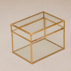 May include: A rectangular glass box with a gold-colored metal frame. The box has a hinged lid and is designed to hold small items. The clear glass allows visibility of the contents. The box is sitting on a neutral-colored surface.
