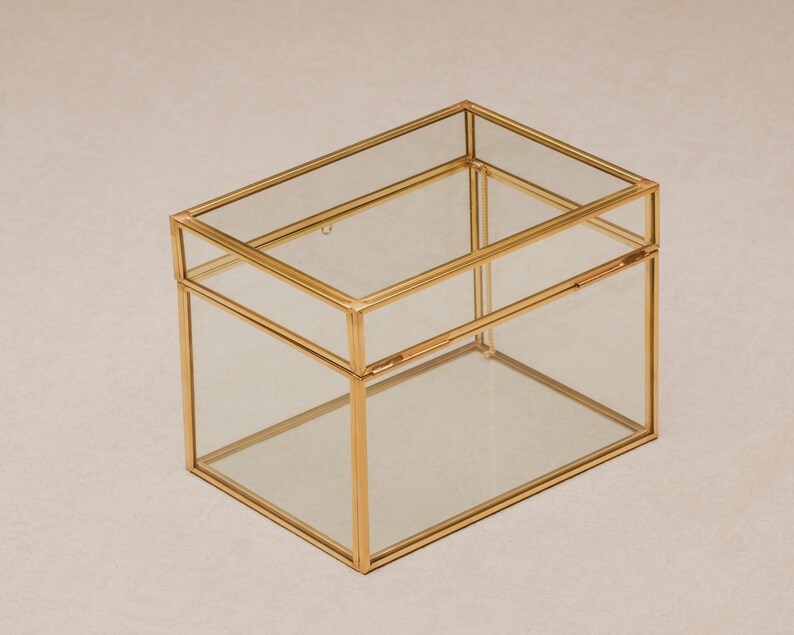 May include: A rectangular glass box with a gold-colored metal frame. The box has a hinged lid and is designed to hold small items. The clear glass allows visibility of the contents. The box is sitting on a neutral-colored surface.