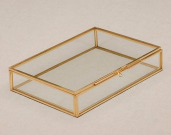 May include: A rectangular glass box with a hinged lid and a gold-colored metal frame. The box is empty and sits on a neutral-colored surface. The lid is slightly ajar, revealing the interior.