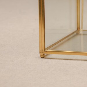May include: A gold-colored, geometric display case with clear glass panels. The case has a cube shape with a delicate, ornate design. The base of the case has small, rounded feet. The frame is made of a shiny, metallic material.