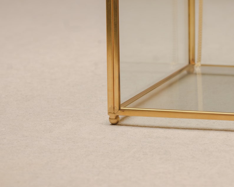 May include: A gold-colored, geometric display case with clear glass panels. The case has a cube shape with a delicate, ornate design. The base of the case has small, rounded feet. The frame is made of a shiny, metallic material.