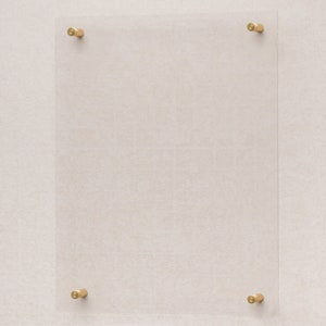 May include: A clear acrylic frame with four gold metal corner brackets. The frame is mounted on a light beige wall.