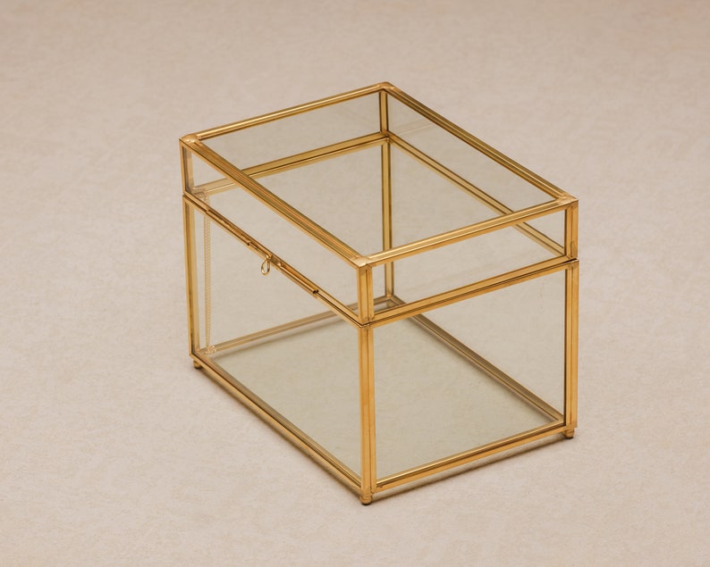 May include: A rectangular glass and gold-tone metal box with a hinged lid. The box has a clear glass body and a gold-colored metal frame. The lid is also glass and has a small latch. The box is sitting on a neutral surface.
