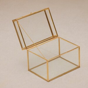 May include: A rectangular glass box with a hinged lid, framed in gold-colored metal. The lid is slightly ajar, revealing the clear interior. The box is designed to display or store small items, offering a modern and elegant aesthetic.