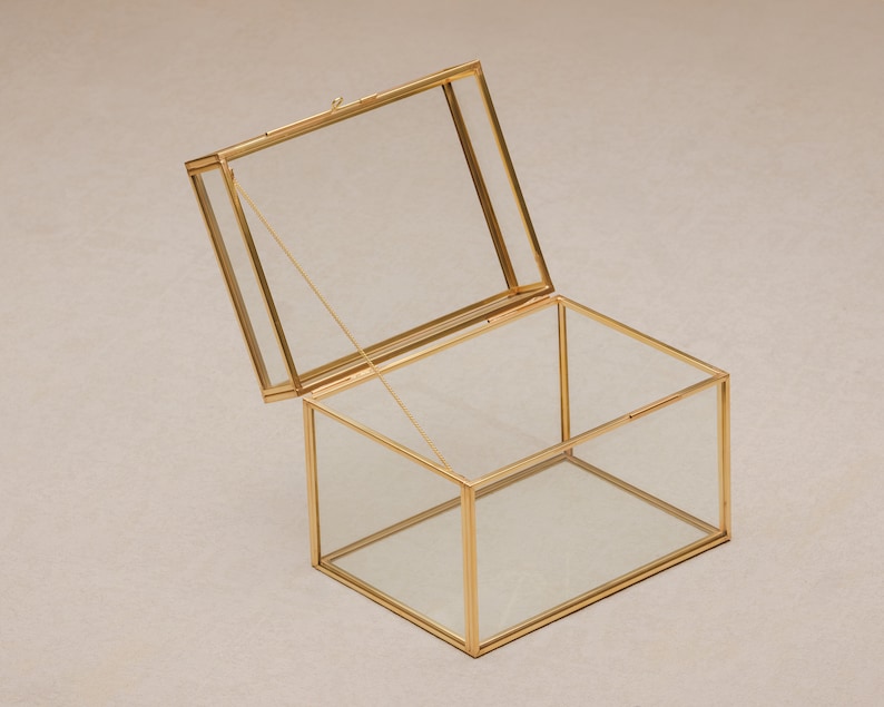 May include: A rectangular glass box with a hinged lid, framed in gold-colored metal. The lid is slightly ajar, revealing the clear interior. The box is designed to display or store small items, offering a modern and elegant aesthetic.