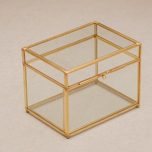May include: A rectangular glass box with a gold-colored metal frame. The box has a hinged lid and a small latch. The clear glass allows visibility of the contents. The box is suitable for storing jewelry or small keepsakes.