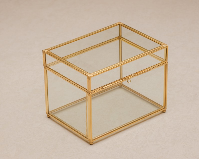 May include: A rectangular glass box with a gold-colored metal frame. The box has a hinged lid and a small latch. The clear glass allows visibility of the contents. The box is suitable for storing jewelry or small keepsakes.