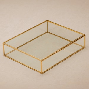 Könnte beinhalten: Eine transparente Glasbox mit goldenem Metallrahmen und einem Scharnierdeckel. Die Box ist leer und hat ein schlichtes Design.