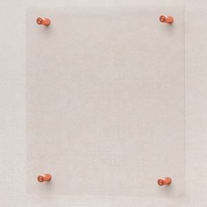 May include: A clear glass frame with four copper-colored mounting brackets. The frame is mounted on a light beige wall.