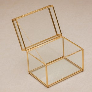 May include: A rectangular glass box with a hinged lid, framed in gold-colored metal. The lid is partially open, revealing the interior. The box is designed to display or store small items. The box is approximately 6 inches long, 4 inches wide, and 3 inches tall.