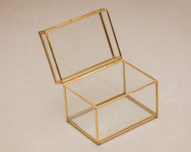 May include: A rectangular glass box with a hinged lid, framed in gold-colored metal. The lid is partially open, revealing the interior. The box is designed to display or store small items. The box is approximately 6 inches long, 4 inches wide, and 3 inches tall.