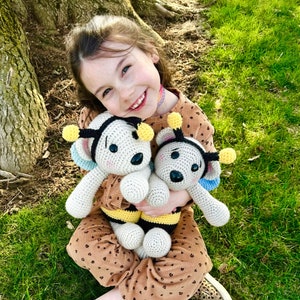 May include: A young girl is sitting on the grass and holding two crocheted bee plushies. The plushies are white with black stripes and yellow accents. The girl is wearing a brown and white patterned dress and brown and white patterned shoes.