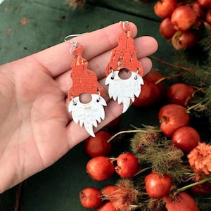 May include: A pair of gnome-shaped earrings. The earrings feature a red cork hat and a white beard. The earrings are held in a hand, with red berries and greenery in the background. The earrings have silver-colored hooks.