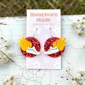 May include: Handmade earrings with a white trainer design and yellow wings, set on a red glitter disc. The earrings are displayed on a pink card with the text "Hometown Made DESIGN COMPANY".