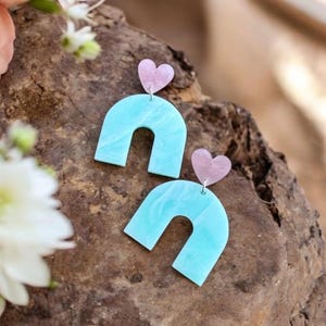 May include: A pair of turquoise blue arch-shaped earrings with pink heart accents. The earrings are made of resin and have a marbled effect.