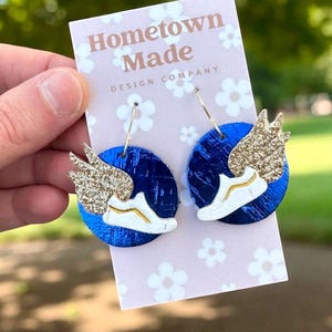 May include: A pair of earrings featuring a blue circular base with a white sneaker design and gold glitter wings. The earrings are attached to gold hoops. The background includes a pink card with the text "Hometown Made DESIGN COMPANY".