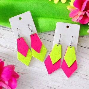 May include: A pair of neon pink and green geometric earrings. The earrings are made of two layers of geometric shapes, one pink and one green. The earrings are hanging from silver hooks.