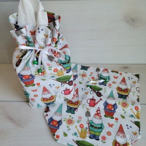 May include: A white fabric tissue holder and matching table runner featuring a whimsical garden gnome print. The gnomes are depicted with red hats, blue jackets, and white beards, surrounded by garden tools and flowers. The tissue holder is gathered at the top with a white ribbon.