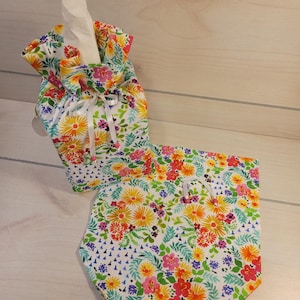 May include: A floral tissue box cover and matching hexagonal tissue holder. The fabric features a white background with a vibrant pattern of yellow, orange, pink, and purple flowers, accented by green leaves. The tissue box cover has a drawstring closure.
