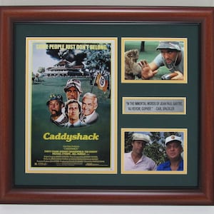 May include: Framed movie poster and stills from the film "Caddyshack." The poster features the film's title and cast, with additional images of scenes and quotes. The frame is dark brown with a green matting.