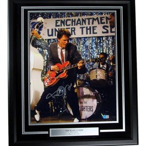 May include: A black and white photo of Michael J. Fox playing guitar on stage with a band. The photo is signed by Michael J. Fox and the band, "Berry and the Lighters". The photo is framed in a black frame with a silver mat.