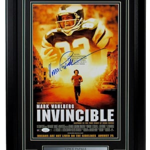 May include: A movie poster for the film "Invincible" starring Mark Wahlberg. The poster features a photo of Wahlberg running down a street, with a football player in the background. The text on the poster reads "Invincible" and "Dreams are not lived on the sidelines." The poster is framed in black and has a silver plaque with the text "Vince Papale, Philadelphia Eagles 1976-1979".