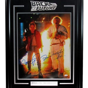 May include: Framed print featuring the "Back to the Future" logo and a movie scene with Michael J. Fox and Christopher Lloyd. The print is in a black frame with white matting, and includes the actors' signatures.