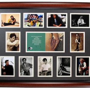 May include: A framed collage featuring multiple photographs of a musician, including a signed album cover. The frame is dark brown with a black matting. The photos show the musician playing guitar and in various poses. The album cover has a green and gold color scheme.