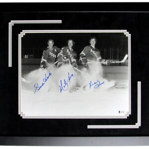 May include: Black and white framed photograph of three hockey players on the ice. The players are wearing jerseys and holding hockey sticks. The photograph is signed in blue ink. The frame is black with a white and gray border.