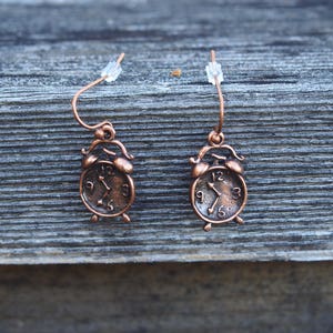 May include: A pair of copper-colored earrings shaped like alarm clocks. The clocks have black hands and Roman numeral markings.