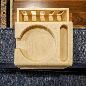 Puede incluir: Una bandeja de brazo de sof&aacute; de madera clara con un portavasos y una ranura para un mando a distancia. La bandeja tiene forma rectangular con una secci&oacute;n elevada en la parte superior y est&aacute; dise&ntilde;ada para colocarse en un brazo de sof&aacute;.