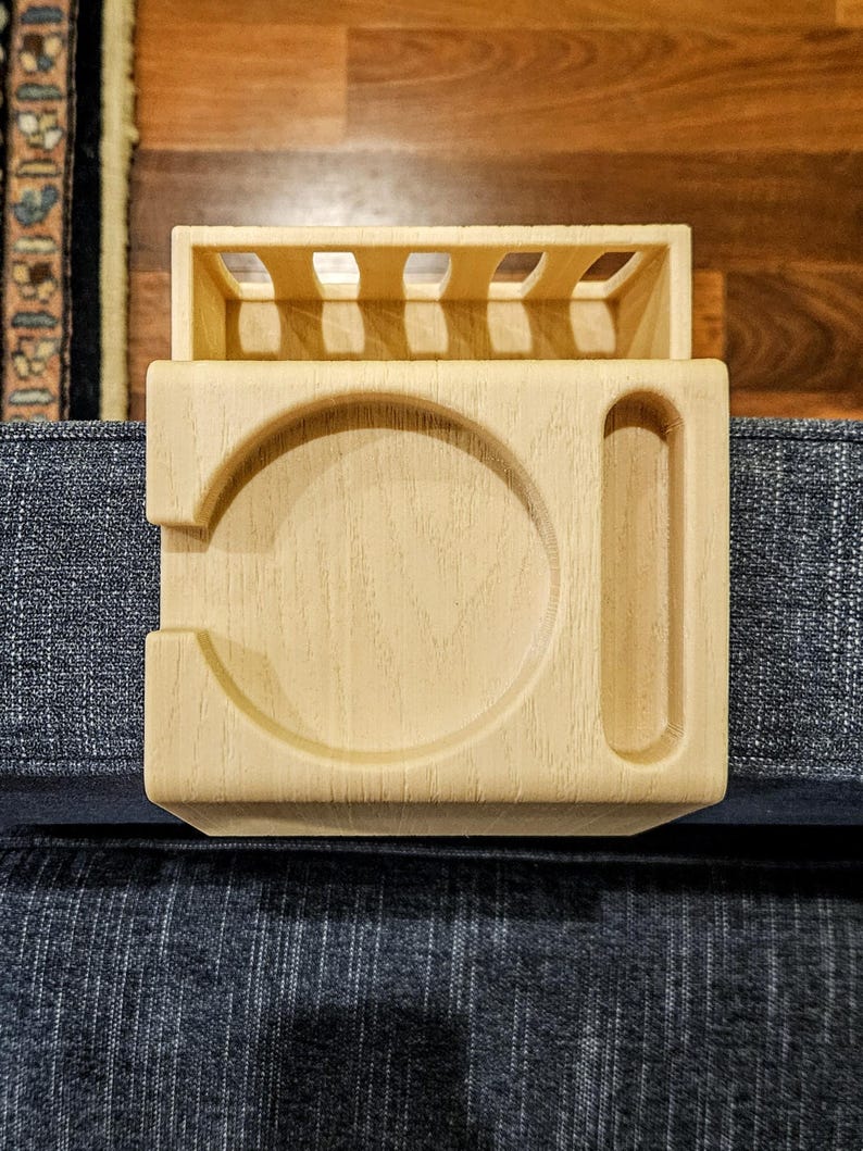 Puede incluir: Una bandeja de brazo de sof&aacute; de madera clara con un portavasos y una ranura para un mando a distancia. La bandeja tiene forma rectangular con una secci&oacute;n elevada en la parte superior y est&aacute; dise&ntilde;ada para colocarse en un brazo de sof&aacute;.