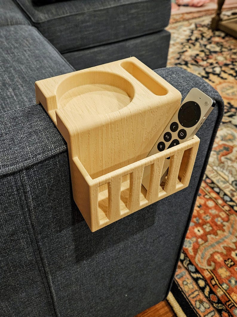 Puede incluir: Un organizador de reposabrazos de sof&aacute; de madera de color claro con un portavasos, una peque&ntilde;a ranura rectangular y un compartimento rectangular para un mando a distancia. El mando a distancia es plateado y negro.