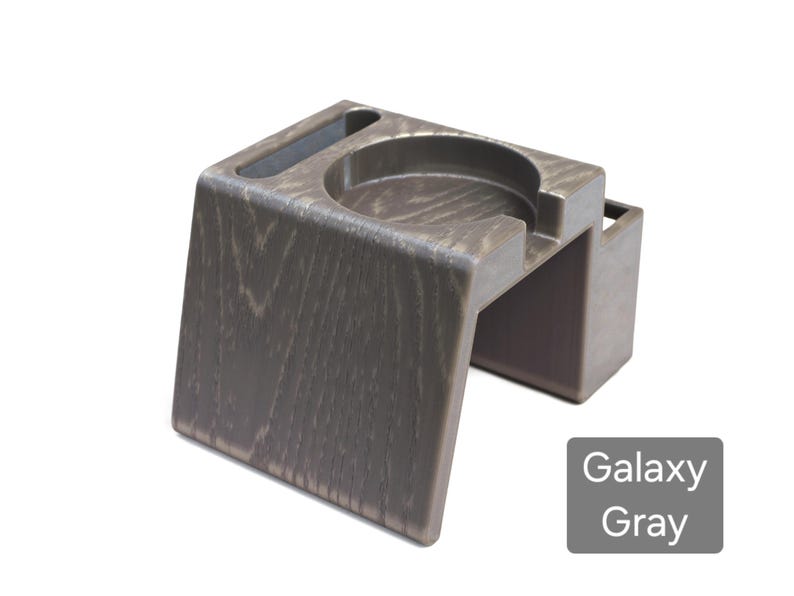 Puede incluir: Un portavasos gris galaxia con una apariencia de vetas de madera texturizada. El portavasos tiene una abertura circular para una taza, una ranura para un tel&eacute;fono y una peque&ntilde;a abertura rectangular. El producto est&aacute; hecho de un material duradero.