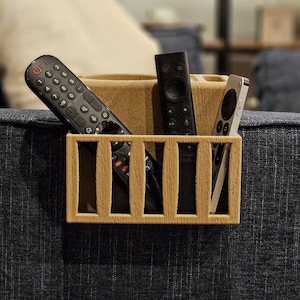 Puede incluir: Un organizador de control remoto de madera con un dise&ntilde;o rectangular y ranuras verticales. El organizador contiene varios controles remotos negros y un control remoto plateado. El organizador se coloca sobre una superficie tapizada azul.