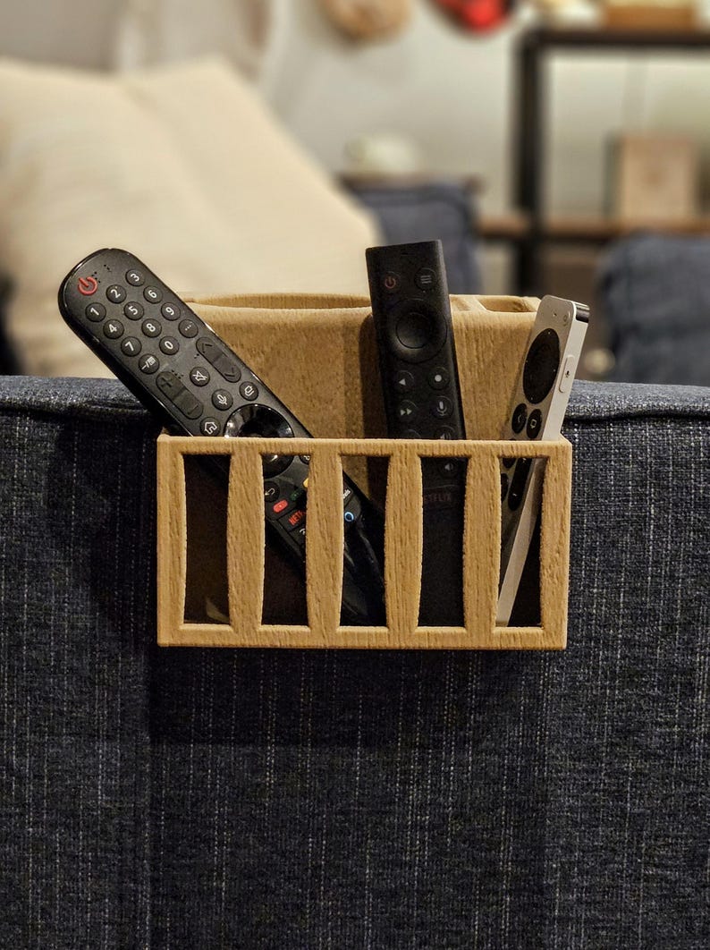 Puede incluir: Un organizador de control remoto de madera con un dise&ntilde;o rectangular y ranuras verticales. El organizador contiene varios controles remotos negros y un control remoto plateado. El organizador se coloca sobre una superficie tapizada azul.