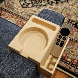 Puede incluir: Una bandeja de brazo de sof&aacute; de madera de color claro con un portavasos circular, un compartimento rectangular y una peque&ntilde;a ranura rectangular. Un mando a distancia plateado descansa en el compartimento rectangular. La bandeja est&aacute; colocada sobre un brazo de sof&aacute; azul.