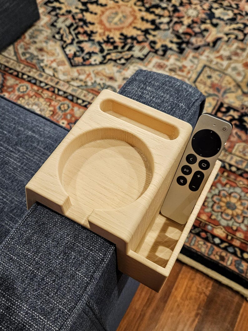 Puede incluir: Una bandeja de brazo de sof&aacute; de madera de color claro con un portavasos circular, un compartimento rectangular y una peque&ntilde;a ranura rectangular. Un mando a distancia plateado descansa en el compartimento rectangular. La bandeja est&aacute; colocada sobre un brazo de sof&aacute; azul.