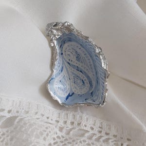 May include: A decorative oyster shell dish with a metallic silver painted exterior edge. The interior of the shell features a light blue base colour with a white swirling paisley pattern and small patches of dark blue paint accents throughout.